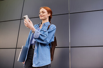 Red haired woman smiles while holding her smartphone in a modern urban setting