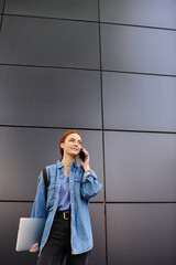 Confident red haired woman chatting on phone while standing in urban setting at midday