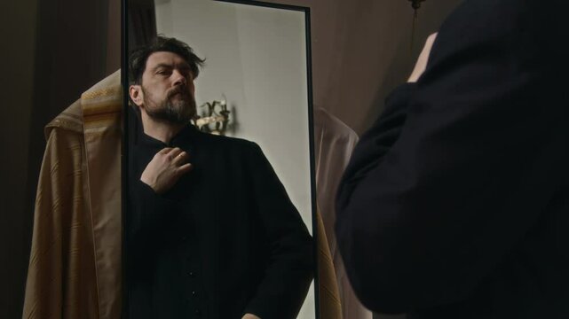 Reflection in mirror of middle-aged Caucasian clergyman adjusting hair and trying on black soutane preparing for religious service in church