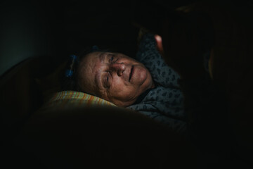 A senior woman lying in bed at night reads a smart phone, her face illuminated by its glow in a...