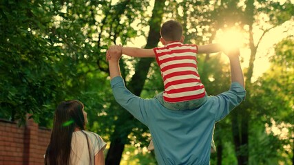 Family walk in park. Child, boy on dad shoulders, spread arms apart, play pilot on background of green trees. Mom, father, son relax in nature in summer. Concept of family leisure. Kid outdoor play - Powered by Adobe