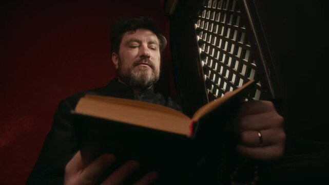Low angle slowmo shot of middle-aged Caucasian priest in black soutane reading Bible out loud through latticed screen to parishioner in confessional booth in church