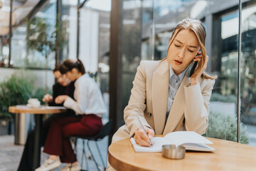 A professional woman speaks on the phone and writes notes at a cafe, conveying focus and multitasking amidst a lively work atmosphere.