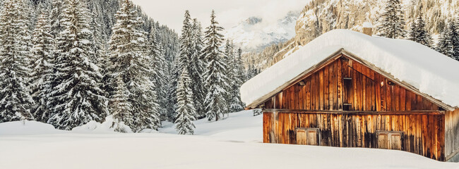 A snow-covered wooden cabin stands in a winter landscape, surrounded by snow-laden pine trees and mountains. San Nicolò Valley, Fassa Valley, Trentino, Italy. 