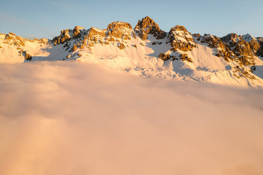 Snow-capped mountains rise above a sea of clouds. Fuciade, Fiamme Valley, Trentino, Italy. Golden light illuminates the peaks and the cloud cover below. - Powered by Adobe
