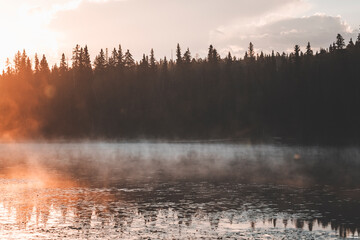 morning hike at sunrise in canada, by a pine tree forest, with misty air and pastel colours, full moon and pale clouds in the sky, by the little quiet pond