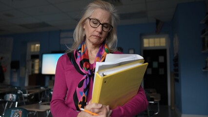 Sad unhappy middle-aged woman in a vibrant sweater holds lesson plans in a well-lit classroom. Her focused expression shows dedication to her students and profession.