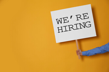 Man holding sign with phrase We Are Hiring on yellow background, closeup. Recruitment, human...