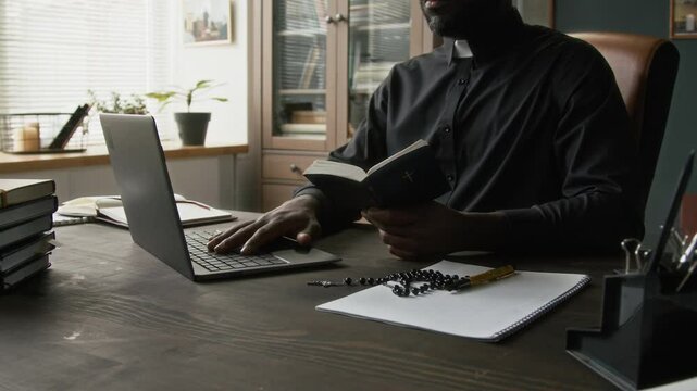 Midsection view of African American young pastor looking at holy bible when preparing slideshow presentation on laptop at large desk in his office