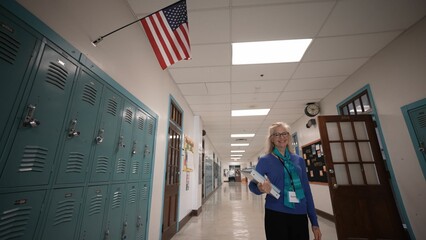 A middle-aged woman teacher stands in a school hallway smiling while holding multiple folders. Lockers line the walls and an American flag hangs overhead creating a welcoming atmosphere.