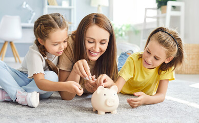 Portrait of happy family of mother and two kids putting money coin into piggy bank lying on floor...