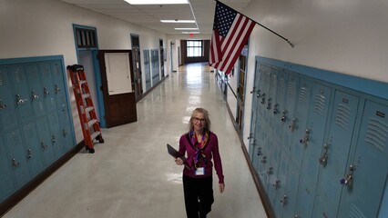 A middle-aged woman teacher with glasses and a purple blouse is walking toward camera in a school hallway holding papers. The hallway features lockers and an American flag hanging above.