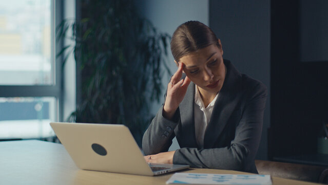 Young businesswoman in office looking stressed while touching her temple and staring at laptop, overwhelmed by workload. Concept of fatigue, pressure, mental strain, and job burnout