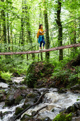 Fototapeta premium Young woman hiker crossing rope bridge over the mountain river in lush forest, wearing yellow backpack and sporty outfit, enjoying outdoor adventure in the wild, active tourism concept