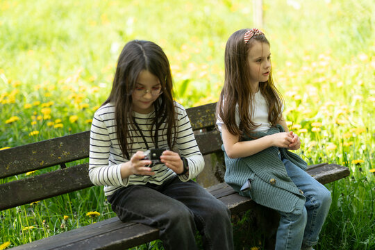 Two setras sit dreamily with a camera in their hands on a wooden bench in a summer park. Concept of children's emotions and leisure