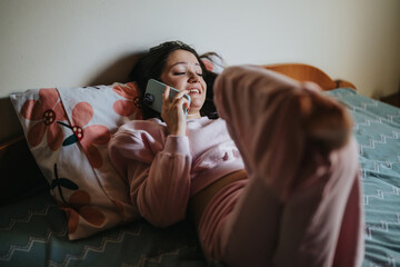 A young woman in casual pink outfit lounging on her bed, smiling while having a phone conversation....