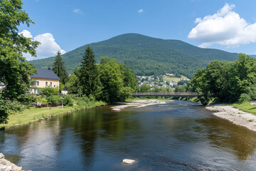 Fototapeta premium Serene river flowing gently beside lush trees and mountains under a clear blue sky on a sunny day