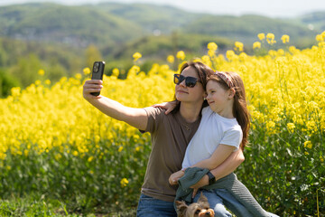 Daughter and mother hugging and smiling taking selfie in front of blooming yellow rapeseed field. Concept of family emotions and leisure