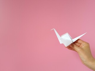 Hand holding white paper crane on pink background. Origami paper crane