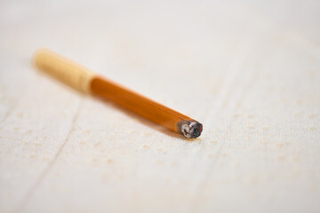 Close-up of lit cigarette resting on light fabric, scattering ash and leaving dark burn mark. Warm tones dominate image, highlighting contrast between burnt and unburnt areas