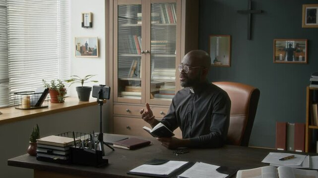 African American young priest having online consultation with parishioner while sitting at office desk holding bible book in front of smartphone mounted on tripod