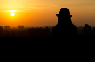 Young woman with long hair and hat gazes at vibrant orange sunset. Silhouette creates serene,...