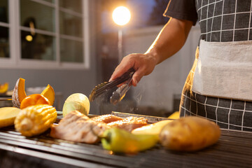 A man enjoys a summer barbecue outdoors, grilling meat and vegetables over open flames, surrounded by friends, smoke, drinks, and weekend freedom.
