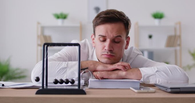 Grumpy tired businessman working with documents in his office, looking at Newton's cradle pendulum, feeling boredom, burnout and dull. Overworked manager having no motivation to continue his work.