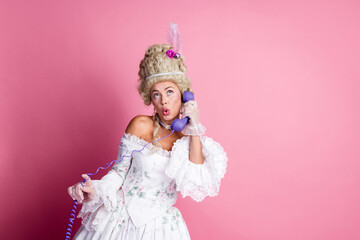 Graceful aristocratic woman in baroque gown holding telephone, posing with humorous expression against pink background