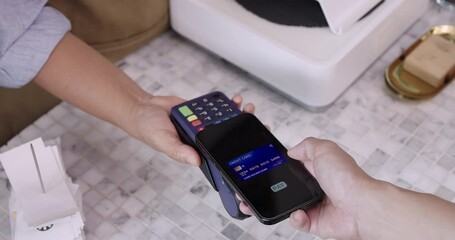 Video of senior Asian female cafe worker receiving digital payment from customer using smartphone and credit card on wireless reader, highlighting contactless transaction in modern cafe scene - Powered by Adobe