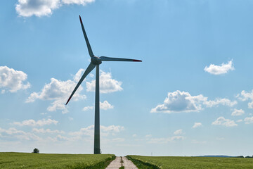 Obraz premium Wind turbine with red tipped blades stands beside cultivated wheat farmland and straight concrete road under clear blue sky evoking balance between human effort and natural resources