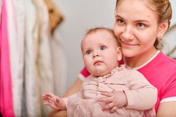Young mother with light brown hair, holding baby girl. Woman wears pink top, baby in light dress. Warm emotions, indoor environment, bright lighting