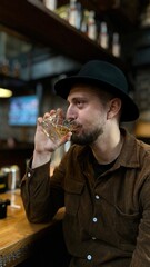Man is drinking whiskey in a pub he is wearing a hat and a brown shirt 