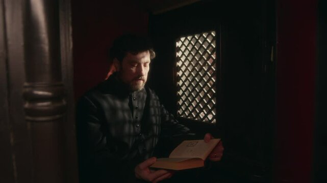 Tilt up shot of middle-aged Caucasian priest in black cassock reading Bible to parishioner in dark confessional booth being illuminated by sunlight coming through latticed screen