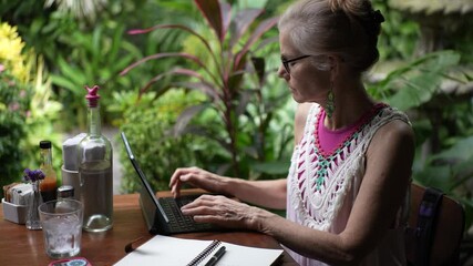 A middle-aged woman sits in a cafe surrounded by lush greenery, focused on her laptop as she balances work and relaxation during her vacation. - Powered by Adobe