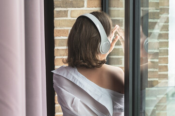 Back view of beautiful сaucasian woman in headphones enjoying listening music standing, leaning back on window.