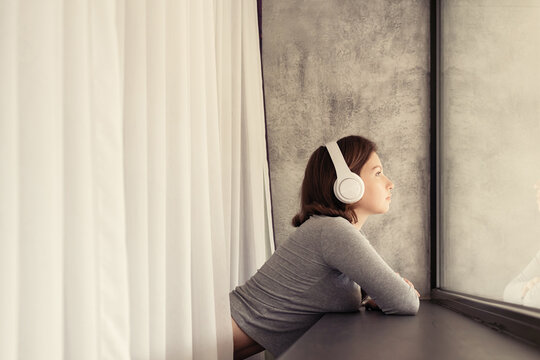 Portrait of beautiful сaucasian young woman in headphones dreaming, enjoying listening music standing and looking near big panoramic window of apartments.