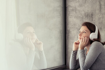 Portrait of beautiful сaucasian young woman in headphones dreaming, enjoying listening music standing near big panoramic window of apartments.