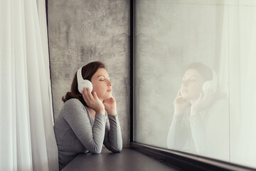 Portrait of beautiful сaucasian young woman in headphones closing eyes dreaming, enjoying listening music standing near big panoramic window of apartments.
