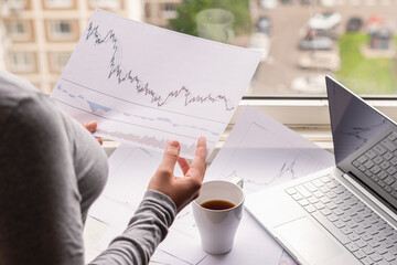 Woman freelancer working on laptop with graphics and charts printed on the paper standing near the window of her apartment. Girl drinking coffee early in the morning. remote work