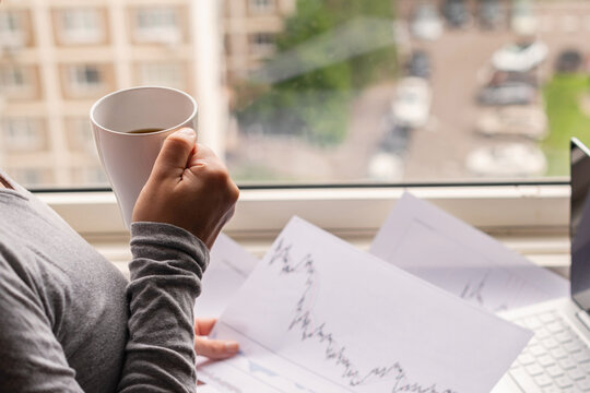 Woman freelancer working on laptop with graphics and charts printed on the paper standing near the window of her apartment. Girl drinking coffee early in the morning. remote work