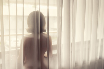 Naked beautiful woman standing behind the transparent curtain near big window.