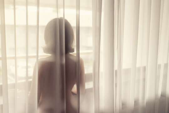 Naked beautiful woman standing behind the transparent curtain near big window.