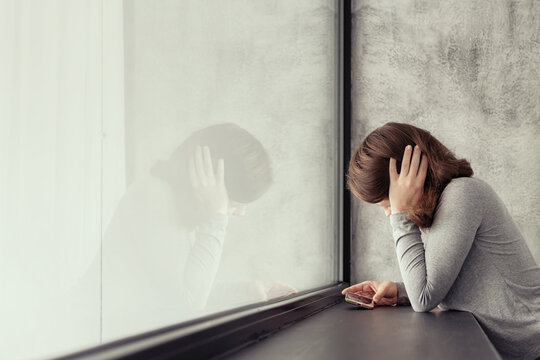 Beautiful сaucasian young woman standing near big panoramic window and looking at mobile phone.