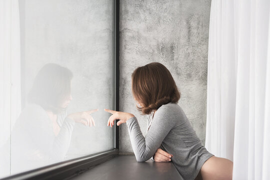 Portrait of beautiful сaucasian young woman touching her reflection of big panoramic window of apartments and listening music.
