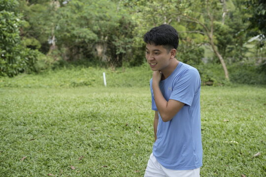 a young man in a light blue shirt stands on a lush green field, surrounded by trees on a sunny day, relaxed and enjoying the outdoors