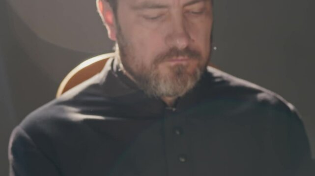 Tilt up close up shot of serious middle-aged Caucasian clergyman in black soutane reading holy book illuminated by sunlight pouring from church window