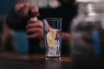 A close-up of a glass being prepared, containing ice cubes and a slice of lemon, against a blurred background emphasizing activity, illustrating beverage preparation at a social bar.