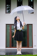 thai university student girl in uniform holding a transparent umbrella while enjoying the rain, standing on a wet reflective pavement in front of a modern thai-style building
