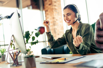 Young businesswoman in a modern office celebrating success while working on a computer wearing a headset with a smile